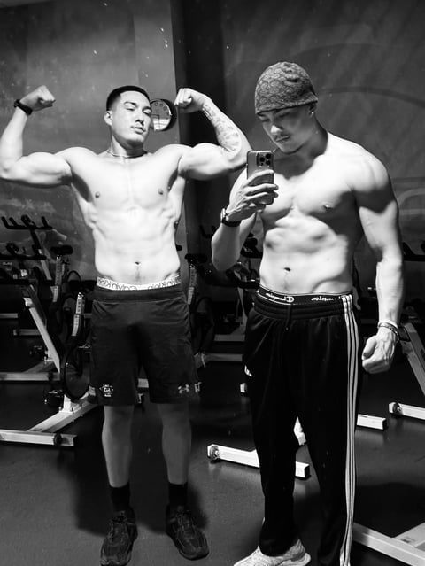 Two shirtless men flexing muscles in a gym, one posing with biceps while the other holds a phone, black and white photo