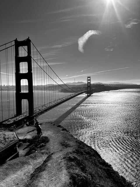 Black and white photograph of the Golden Gate Bridge spanning across San Francisco Bay with mountains in the background and bright sunlight overhead