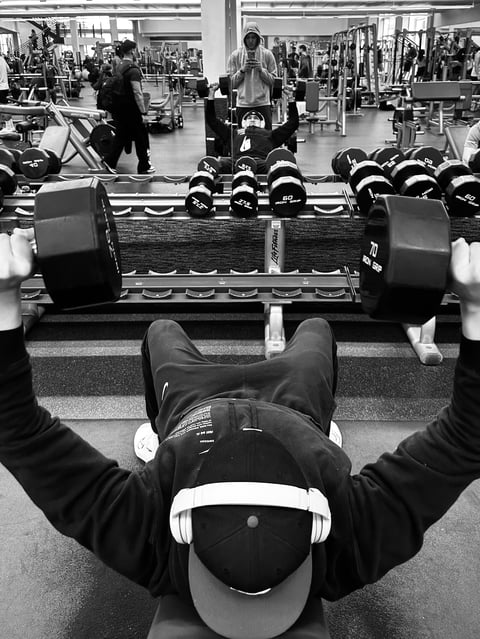 Black and white overhead view of person lifting heavy dumbbells in a gym surrounded by other fitness equipment and gym members